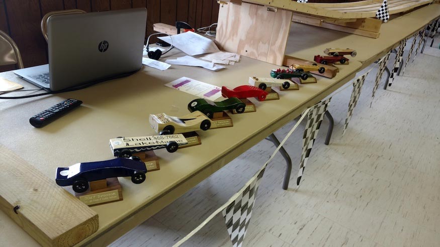 A row of pinewood derby cars lined up on a folding table in front of the racetrack.